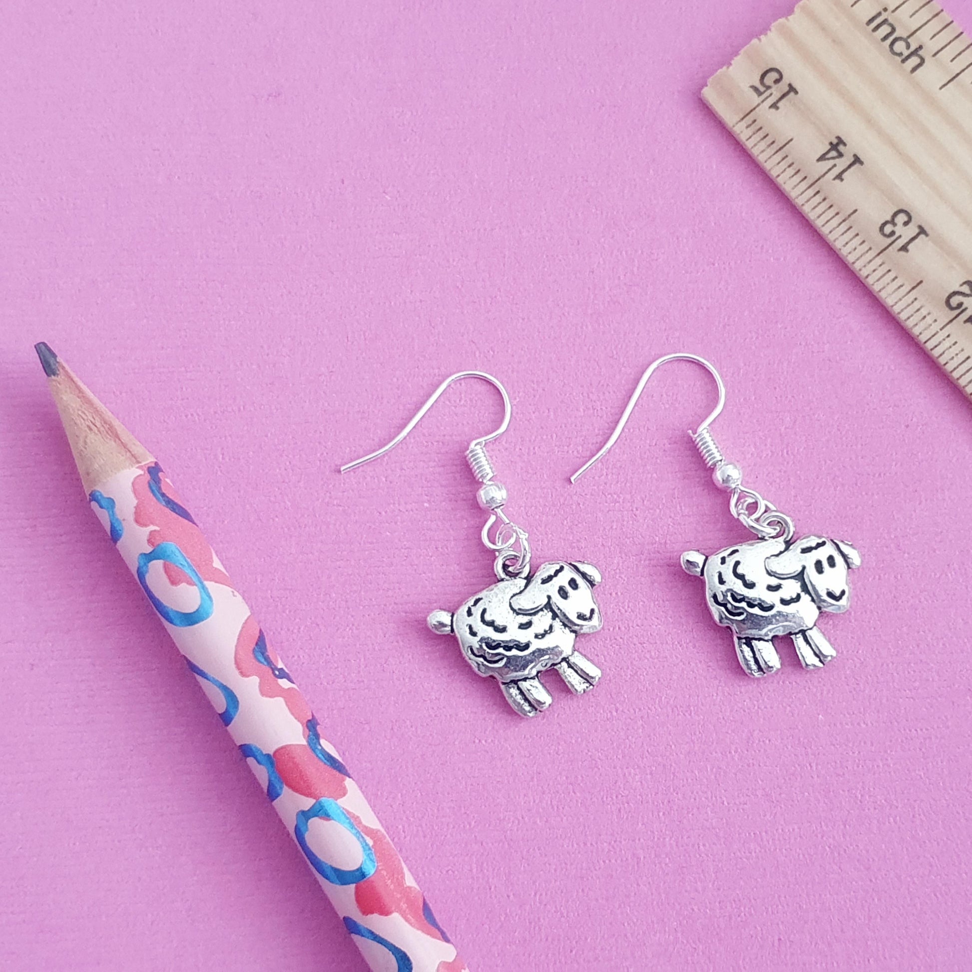 Silver sheep-shaped earrings on a pink background with a ruler and pencil for scale.