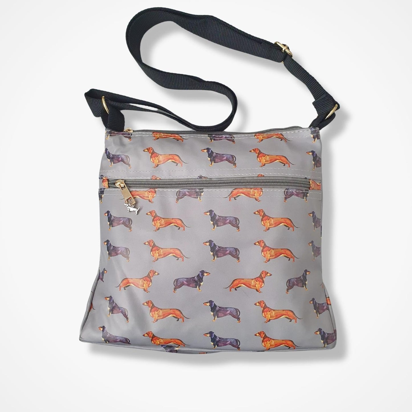 A gray crossbody bag with an allover print of dachshund dogs in orange and brown, featuring a black shoulder strap and a front zippered pocket.