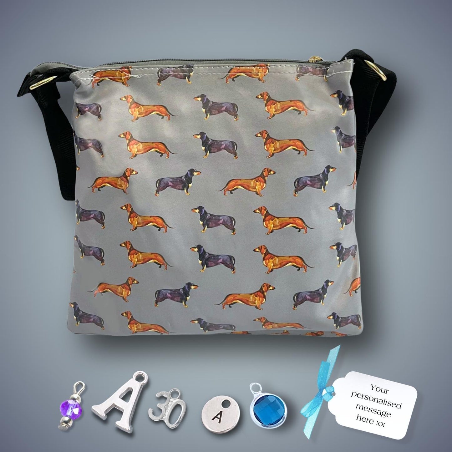 A grey shoulder bag with an allover print of dachshund dogs in orange, black, and purple, featuring black straps and gold-tone hardware. It is displayed on a grey gradient background with customizable charms, initial letters, and a message label shown below it.