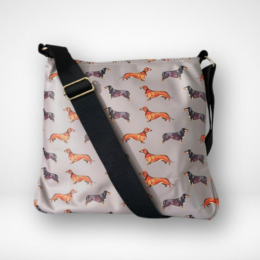 A light gray crossbody bag featuring an all-over print of dachshund dogs in orange, black, and brown, with a black adjustable shoulder strap and a gold-tone buckle.