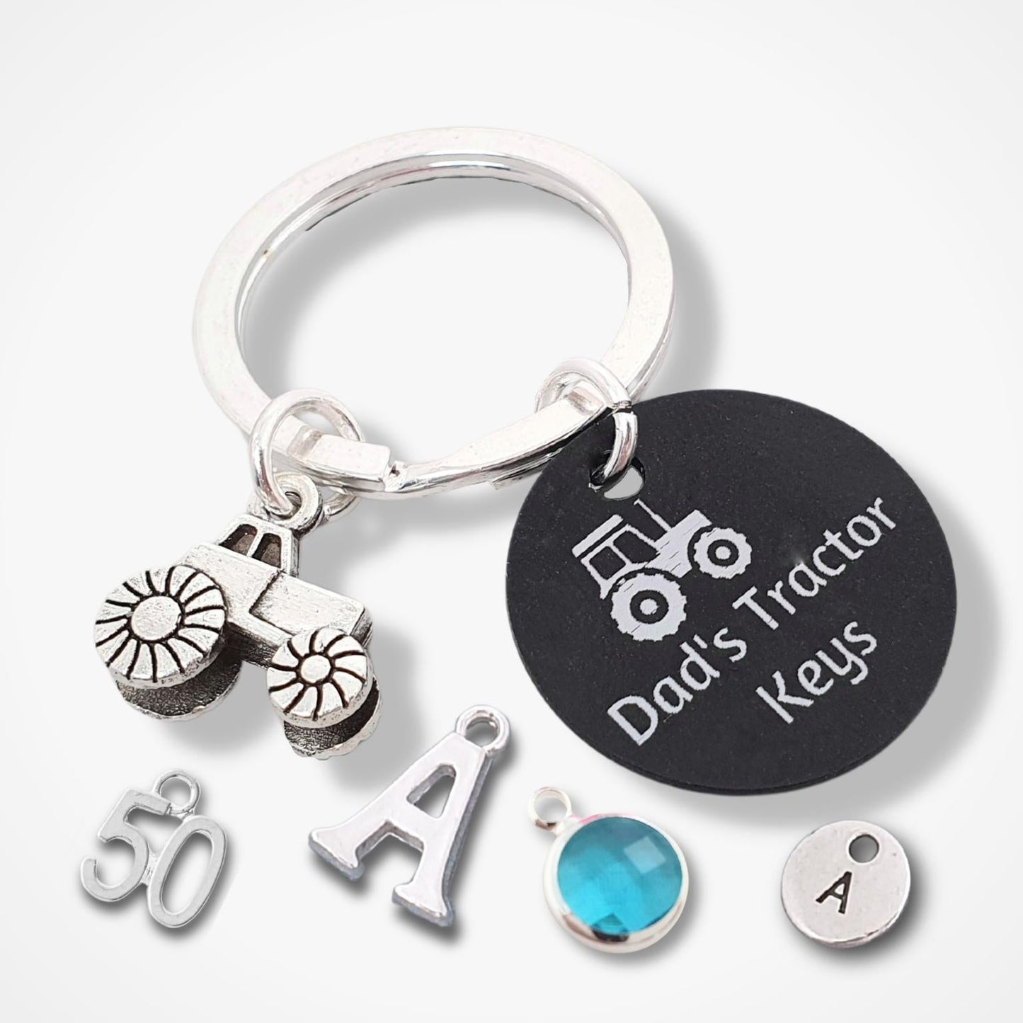A silver tractor-themed keychain with a split ring, featuring a black round tag engraved with 'Dad's Tractor Keys' and small tractor, and letter charms, shown on a light gray background.