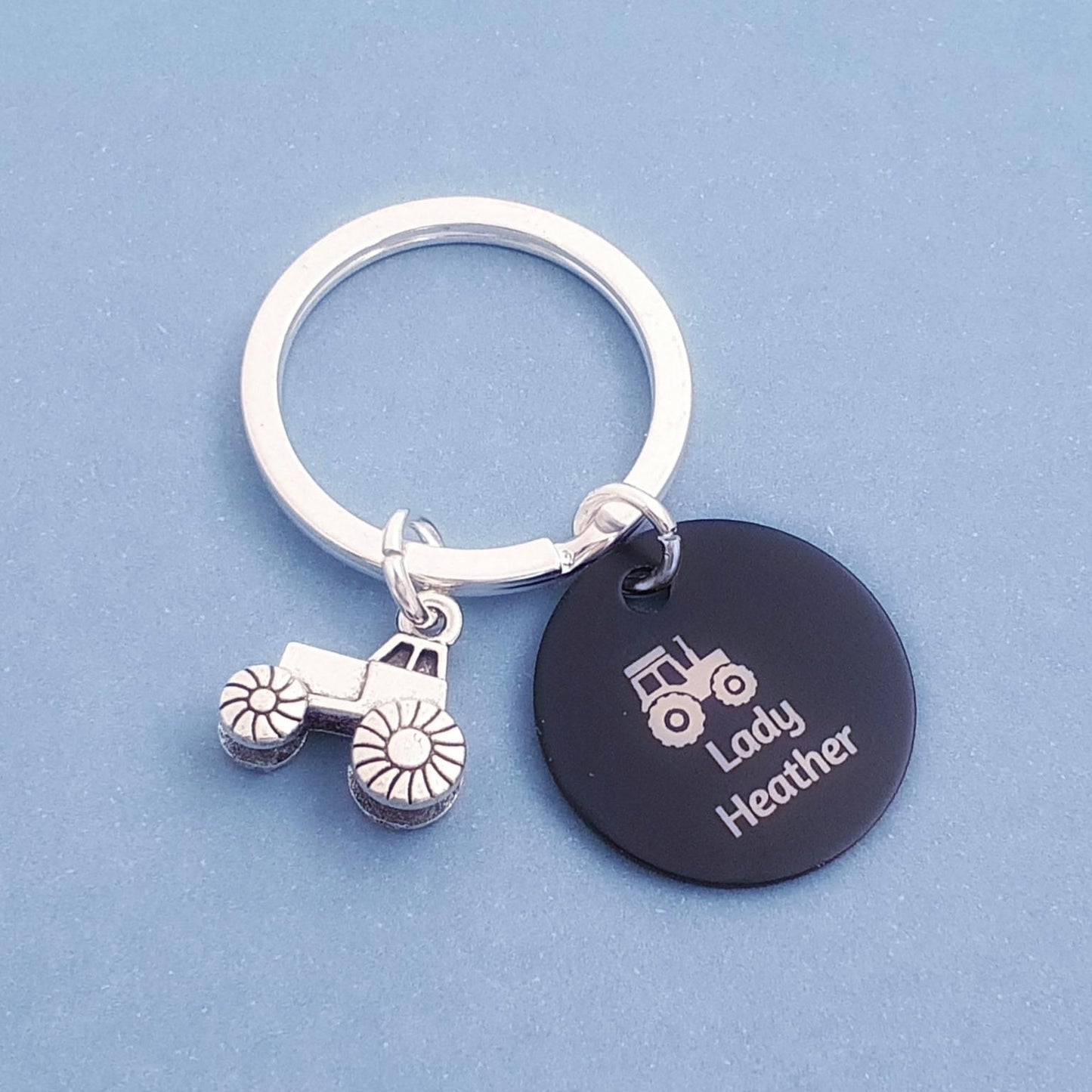 A silver metal keychain with a tractor charm and a round black disc charm engraved with a tractor icon and the names 'Lady Heather', shown on a solid blue background.