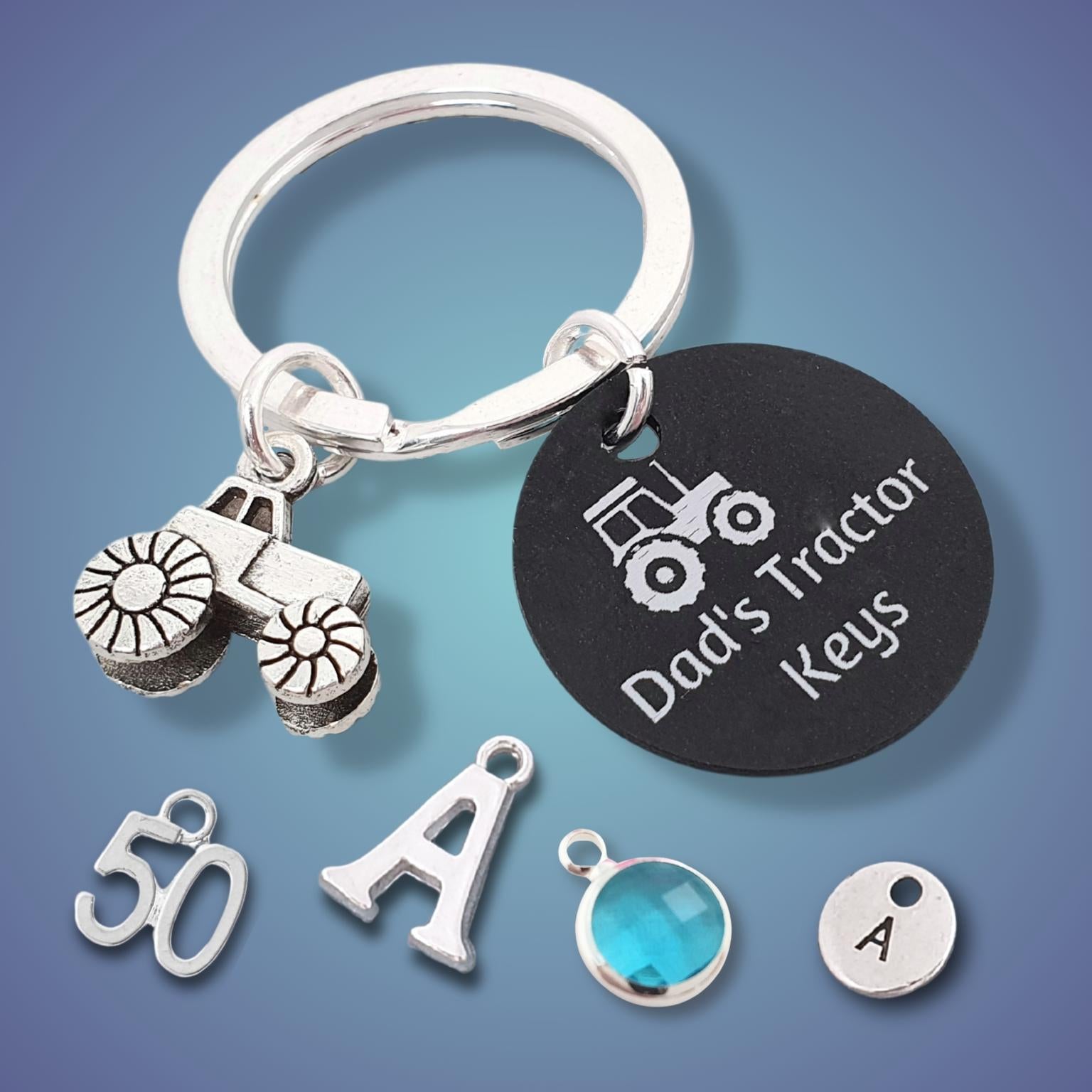 A silver tractor keychain with a black round tag engraved 'Dad's Tractor Keys' and a tractor charm, shown on a blue gradient background with additional customizable charms including a '50' charm, an 'A' letter charm, a blue gem, and a small round 'A' charm.