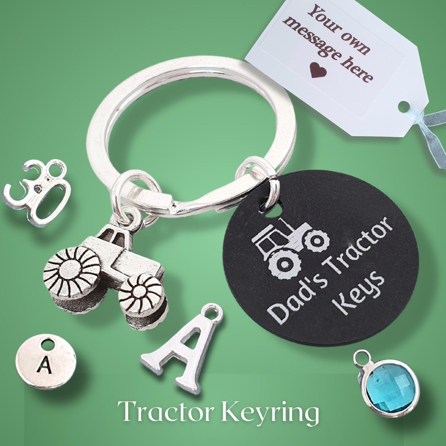 A silver tractor keyring with multiple charms, including a tractor, a letter 'A', a tractor outline, a blue faceted gemstone, and a black round tag engraved with 'Dad's Tractor Keys'. A white tag at the top right says 'Your own message here'.