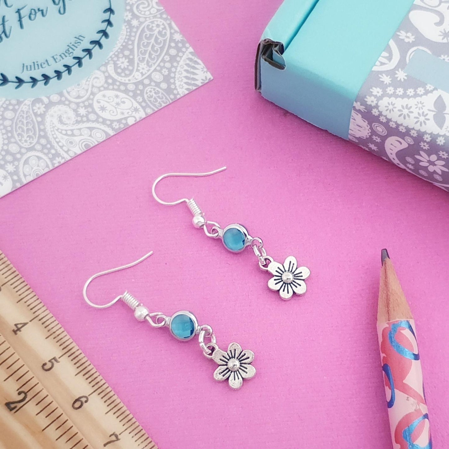 Silver earrings with blue beads and flower charms on a pink surface with a ruler, pencil, and packaging.
