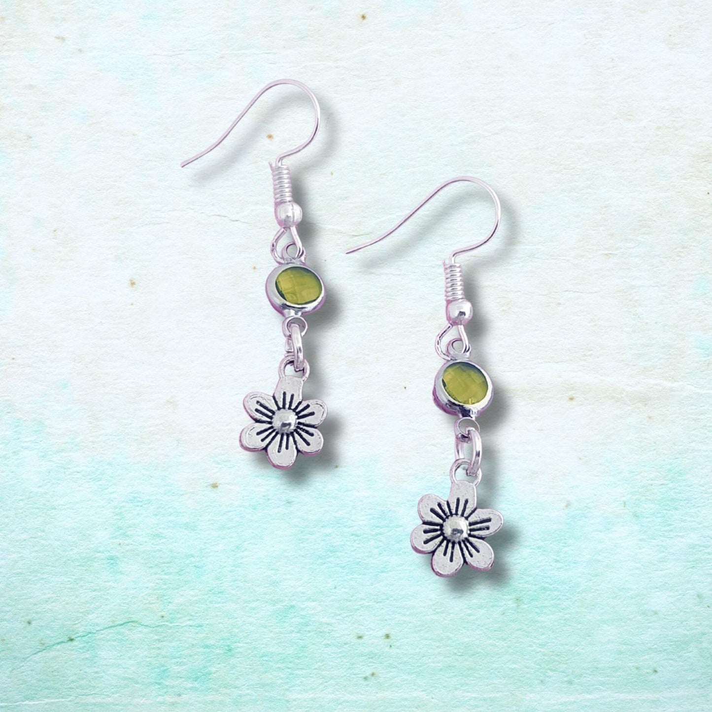 Silver earrings with flower charms and green stones on a light blue textured background