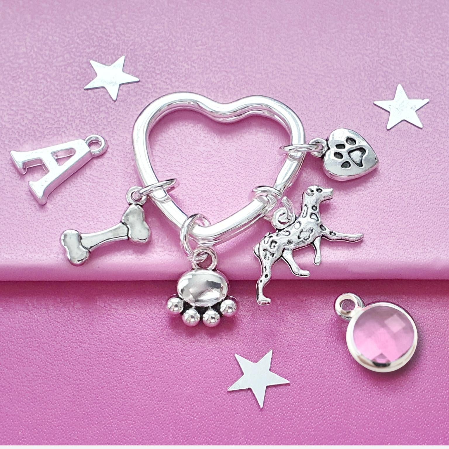 A silver-tone charm keyring with multiple dog-themed charms, including a heart-shaped ring, bone, dog silhouette, paw prints, and a heart with a paw print, displayed on a pink background with star cutouts.