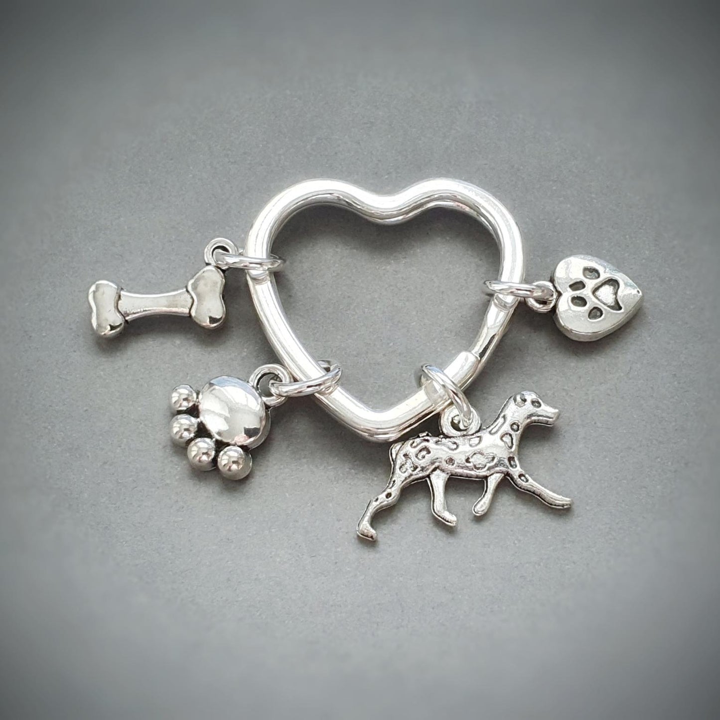 A silver heart-shaped charm keychain with multiple small charms, including a dog silhouette, paw prints, a bone, and a heart, displayed on a dark gray background.