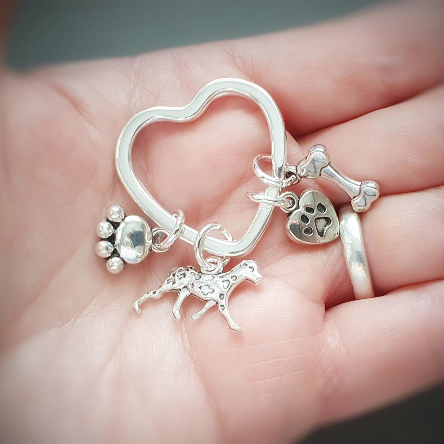 A close-up of a hand holding a silver heart-shaped charm keyring with dog-themed charms, including a paw print, bone, heart, and dog silhouette.