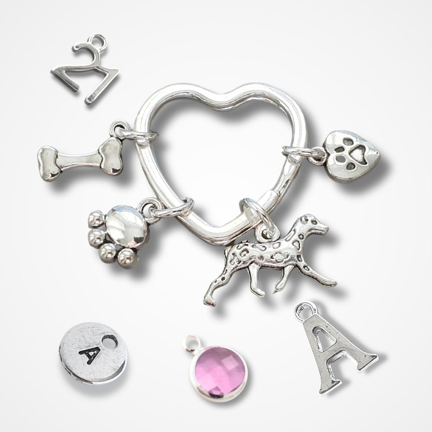A silver pet memorial charm keyring with multiple charms, including a heart-shaped ring, a dog silhouette, paw prints, a bone, and a pink gemstone charm, displayed on a light gray background.