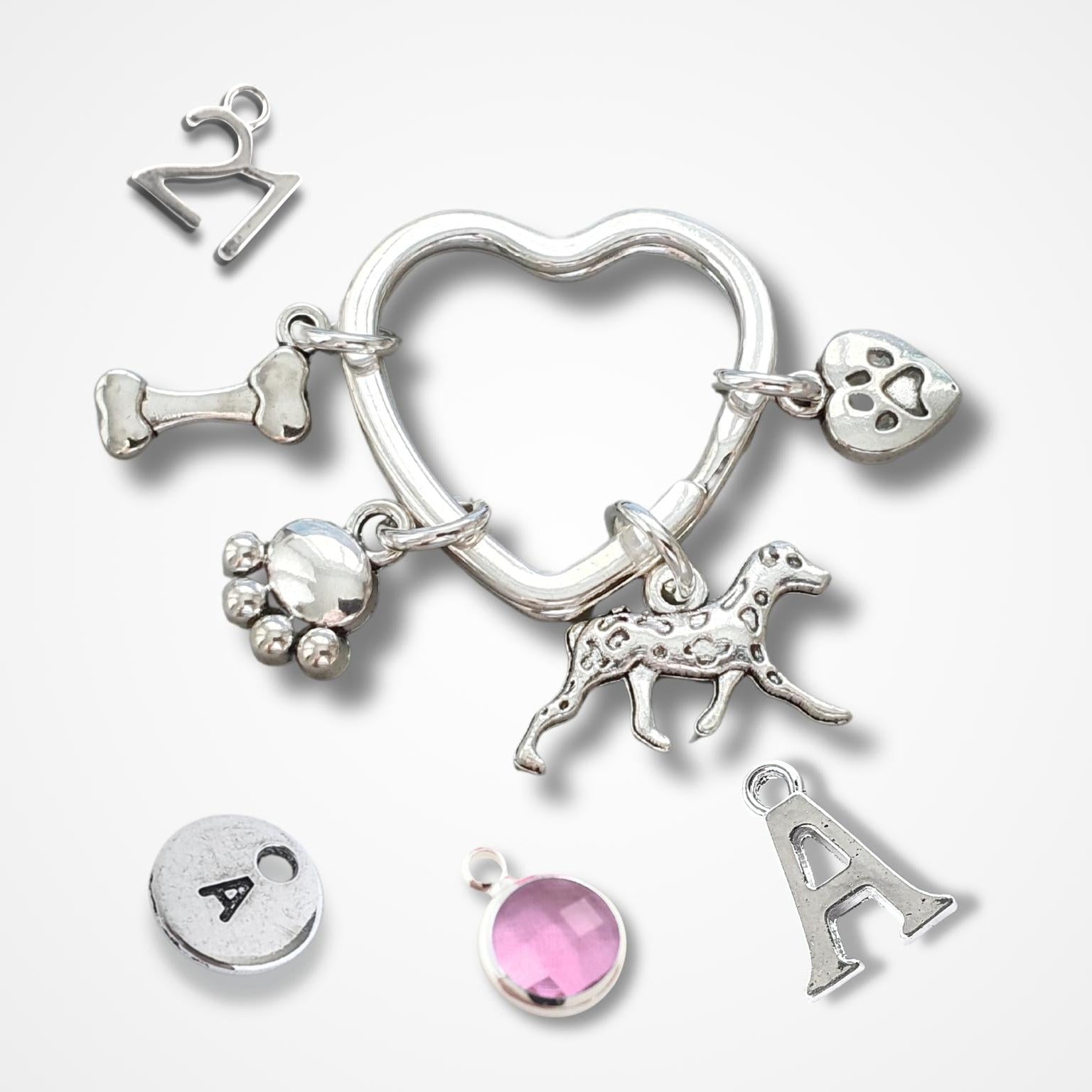 A silver pet memorial charm keyring with multiple charms, including a heart-shaped ring, a dog silhouette, paw prints, a bone, and a pink gemstone charm, displayed on a light gray background.