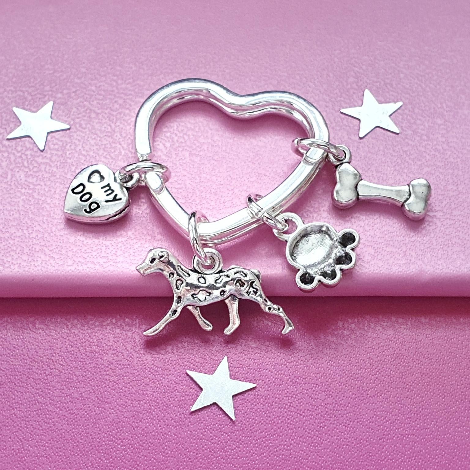 A silver charm bag charm featuring a heart-shaped ring, a dog silhouette charm, a paw print charm, a bone charm, and a small heart charm engraved with '♡ my dog', displayed on a pink background with white star cutouts.