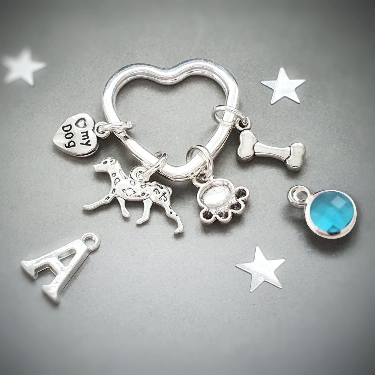 A silver pet memorial key ring charm set featuring a heart-shaped ring, a bone charm, a dog silhouette charm, a paw print charm, a small heart charm engraved with 'My Dog', a letter 'A' charm, and a round blue gemstone charm, displayed on a dark gray background with decorative white star cutouts.