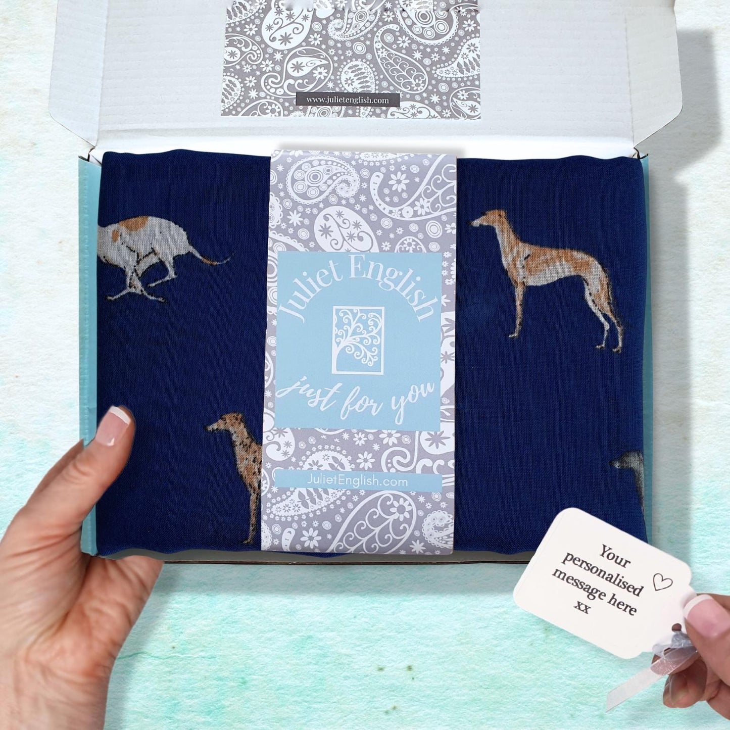 A navy blue scarf with greyhound dog illustrations, presented in an open gift box with a Juliet English band and a personalized message tag held by hands.