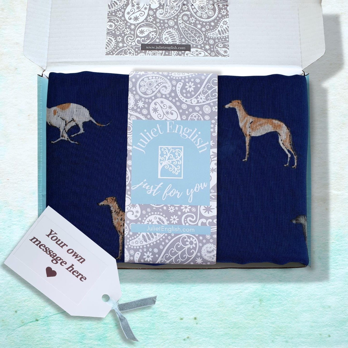 A gift box containing a folded navy blue scarf with dog illustrations, a central white and blue patterned card, and a white gift tag on a light blue textured background.