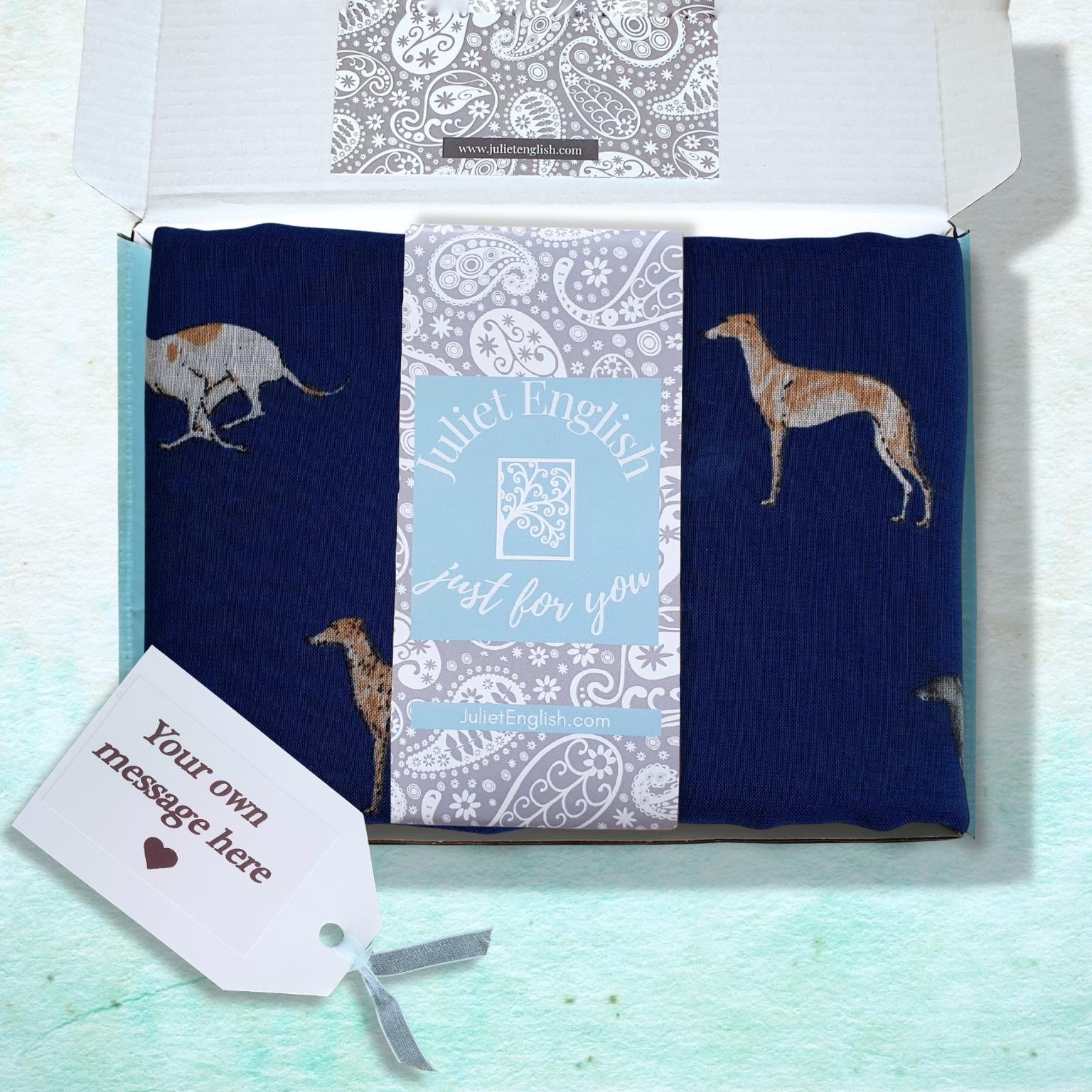A gift box containing a folded navy blue scarf with dog illustrations, a central white and blue patterned card, and a white gift tag on a light blue textured background.
