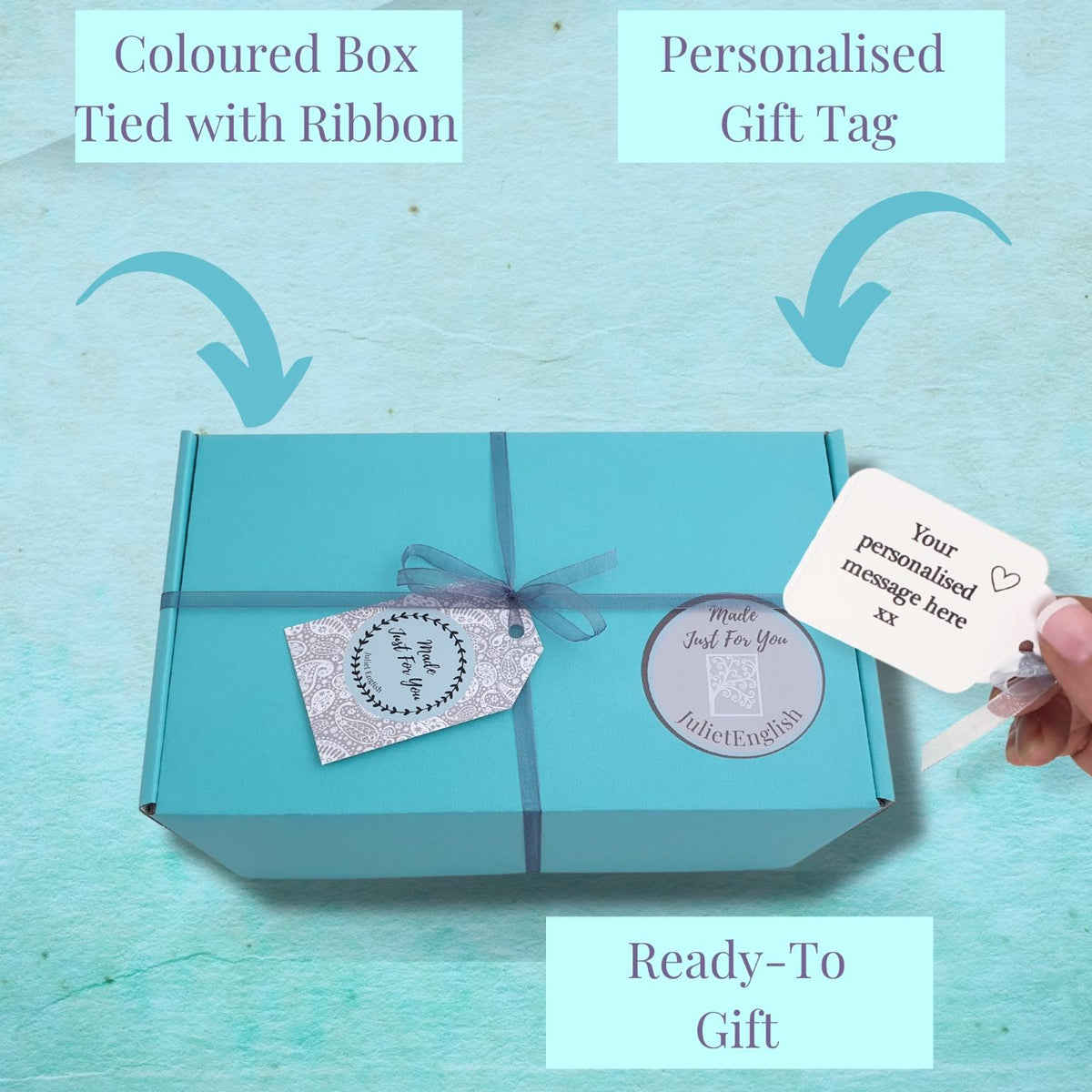 A turquoise gift box tied with a matching ribbon and decorated with two personalized gift tags, shown on a light aqua textured background with callout labels reading 'Coloured Box Tied with Ribbon,' 'Personalised Gift Tag,' and 'Ready-To-Gift.' A hand is holding one of the tags on the right.