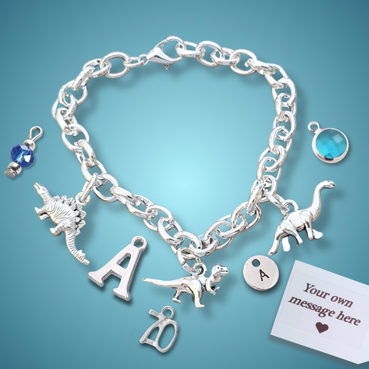 A silver charm bracelet displayed on a blue gradient background. The bracelet features dinosaur charms, letter charms (including 'A' and 'D'), a blue faceted crystal charm, a blue oval stone charm, and a white customizable message tag.
