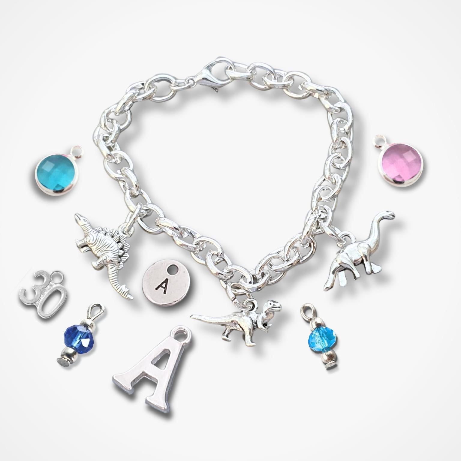 A silver charm bracelet with dinosaur and letter charms, displayed on a clean white background. The bracelet features a silver chain with various charms including three dinosaur figures, a round letter 'A' disc, a '30' charm, a '4' charm, and two round crystal beads in blue and pink.