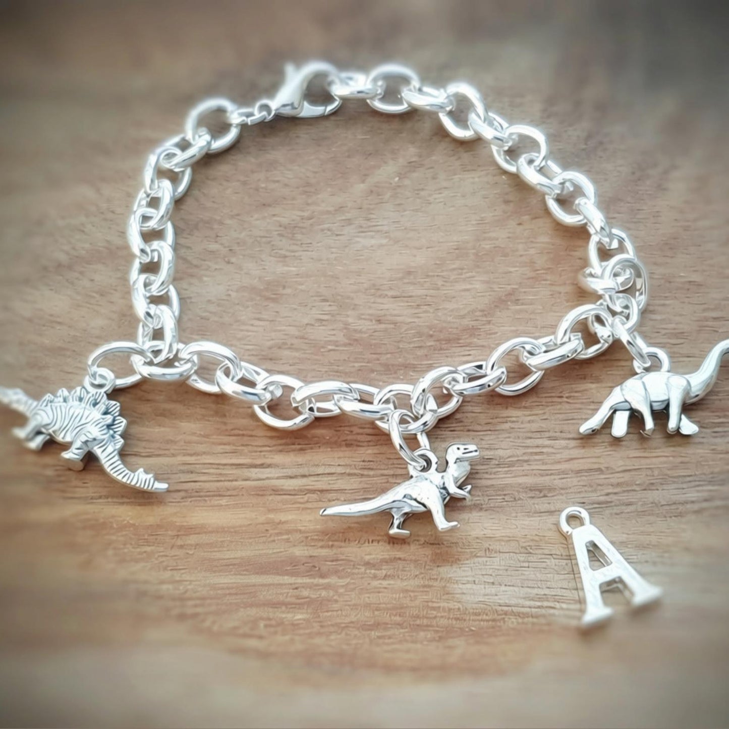 A silver charm bracelet with dinosaur charms and letter 'A' charms, displayed on a wooden surface.