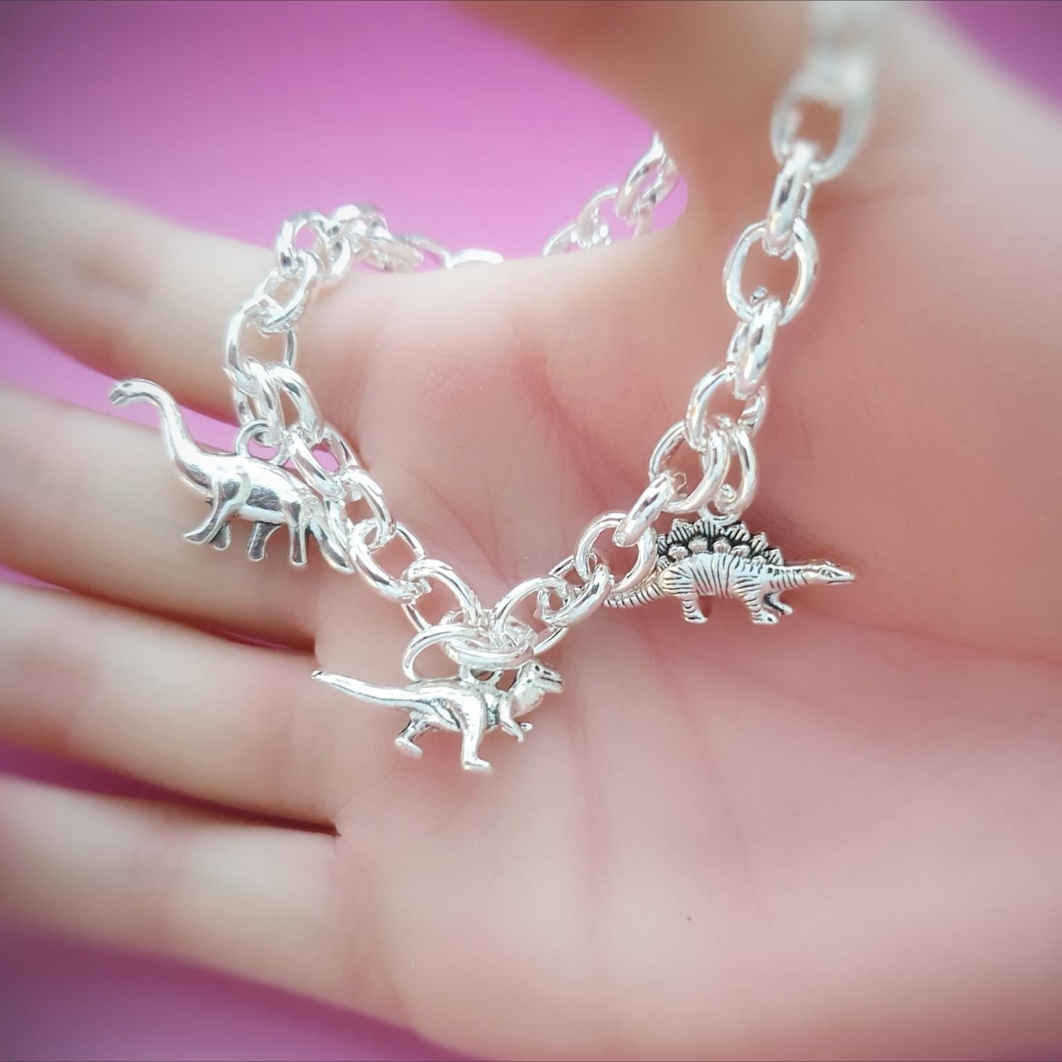 A silver chain bracelet with three dinosaur charms, held in a person's hand against a pink background.