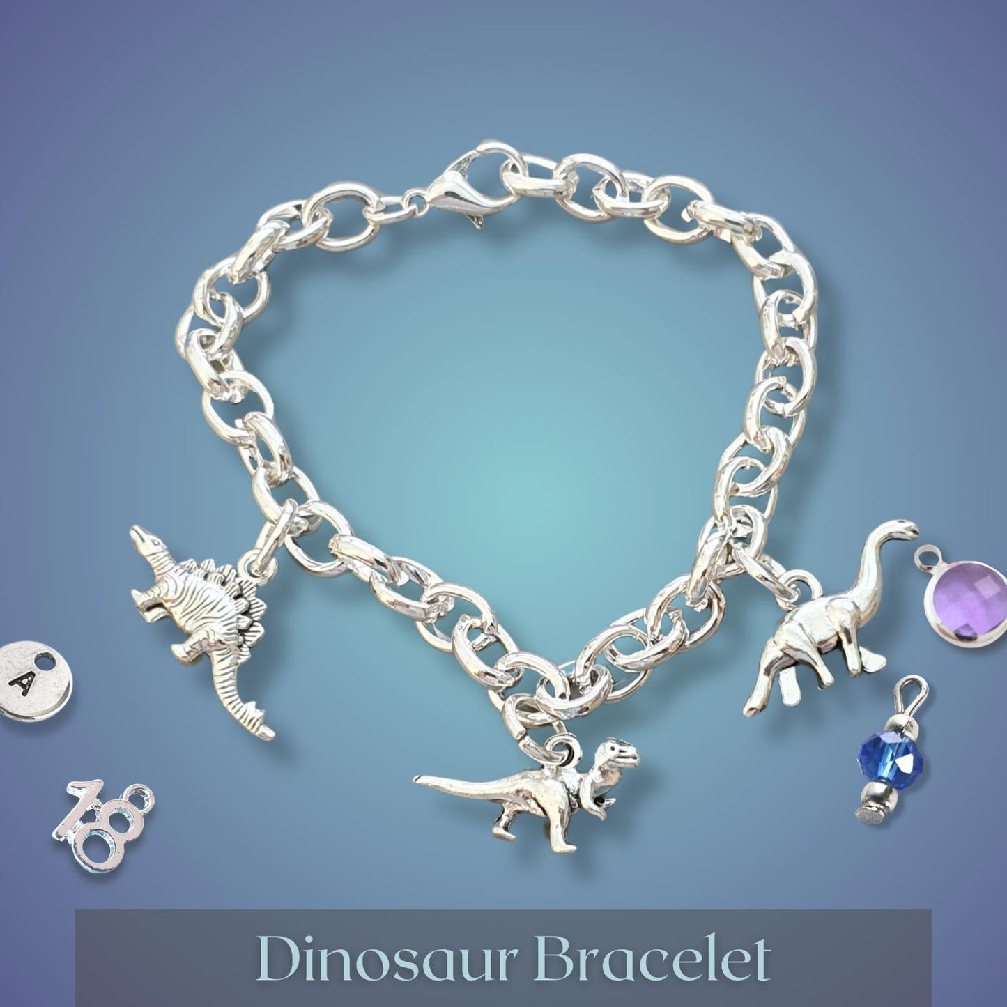 A silver-tone charm bracelet displayed on a blue gradient background, featuring dinosaur charms, a round purple gem, and two loose charm components. The text 'Dinosaur Bracelet' is shown at the bottom.