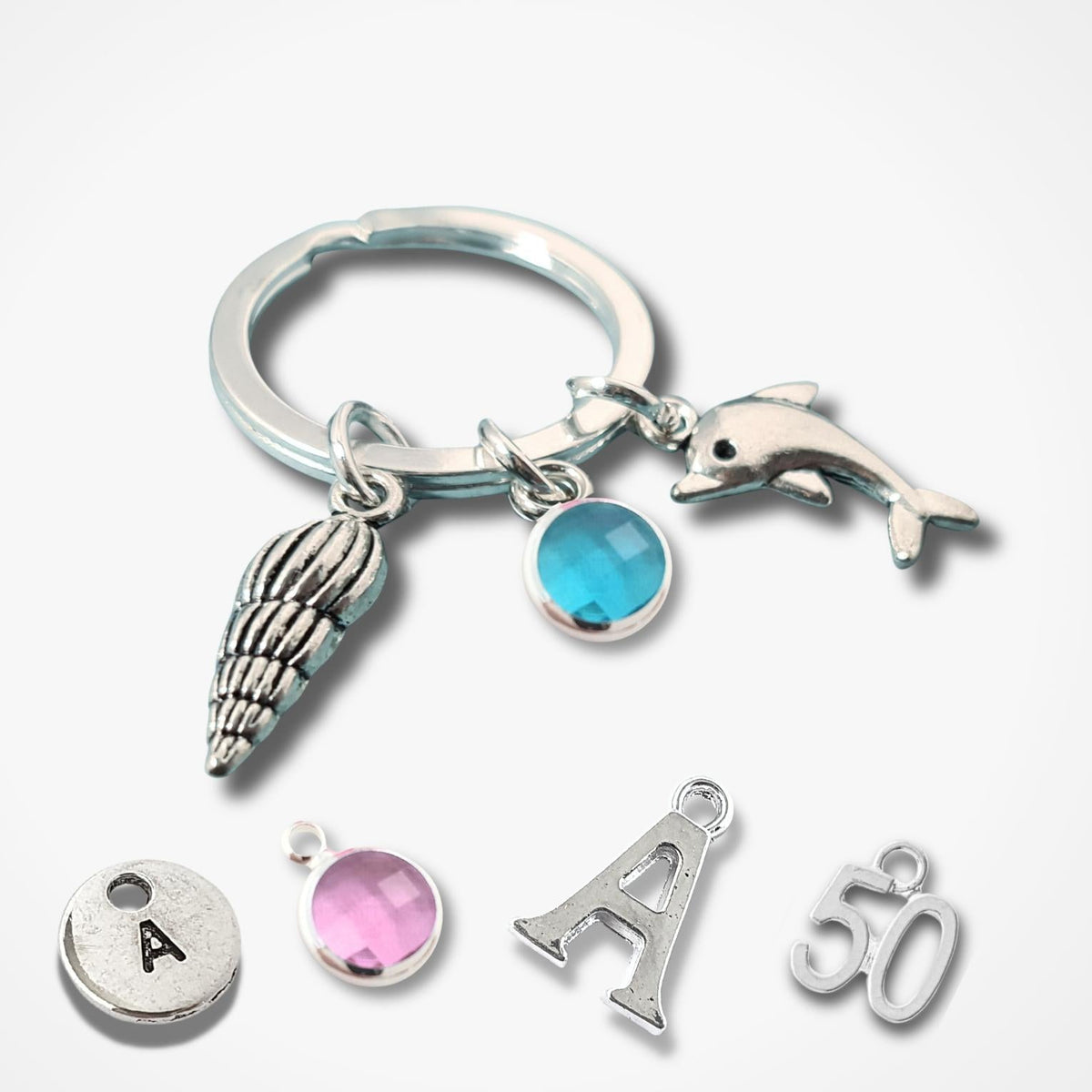 A silver keychain with charms including a dolphin, seashell, and blue stone, shown alongside matching initial 'A' and number '50' charms on a light gray background.