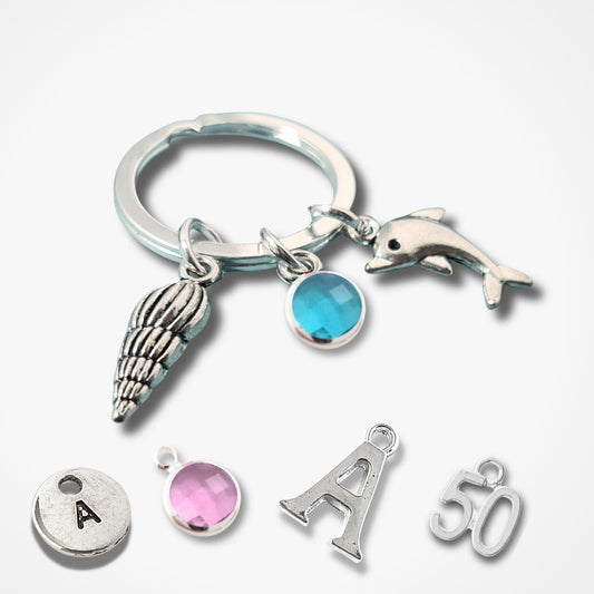 A silver keychain with charms including a dolphin, seashell, and blue stone, shown alongside matching initial 'A' and number '50' charms on a light gray background.