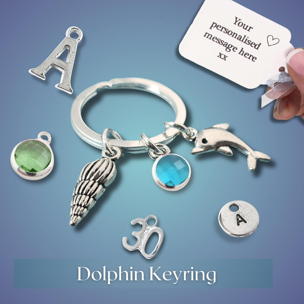 A silver dolphin keyring charm set displayed on a blue gradient background, featuring a dolphin, seashell, letter 'A', '30', and gemstone charms. A hand is holding a white personalization tag, and the text 'Dolphin Keyring' appears at the bottom.