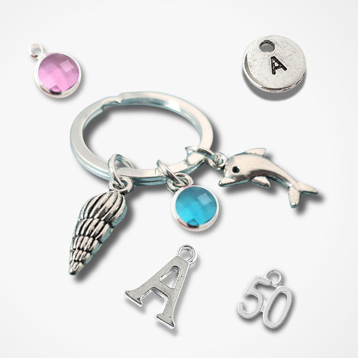 A silver keychain set with charms including a dolphin, shell, letter A, number 50, and two round bezel-set rhinestones in pink and blue, displayed on a clean white background.