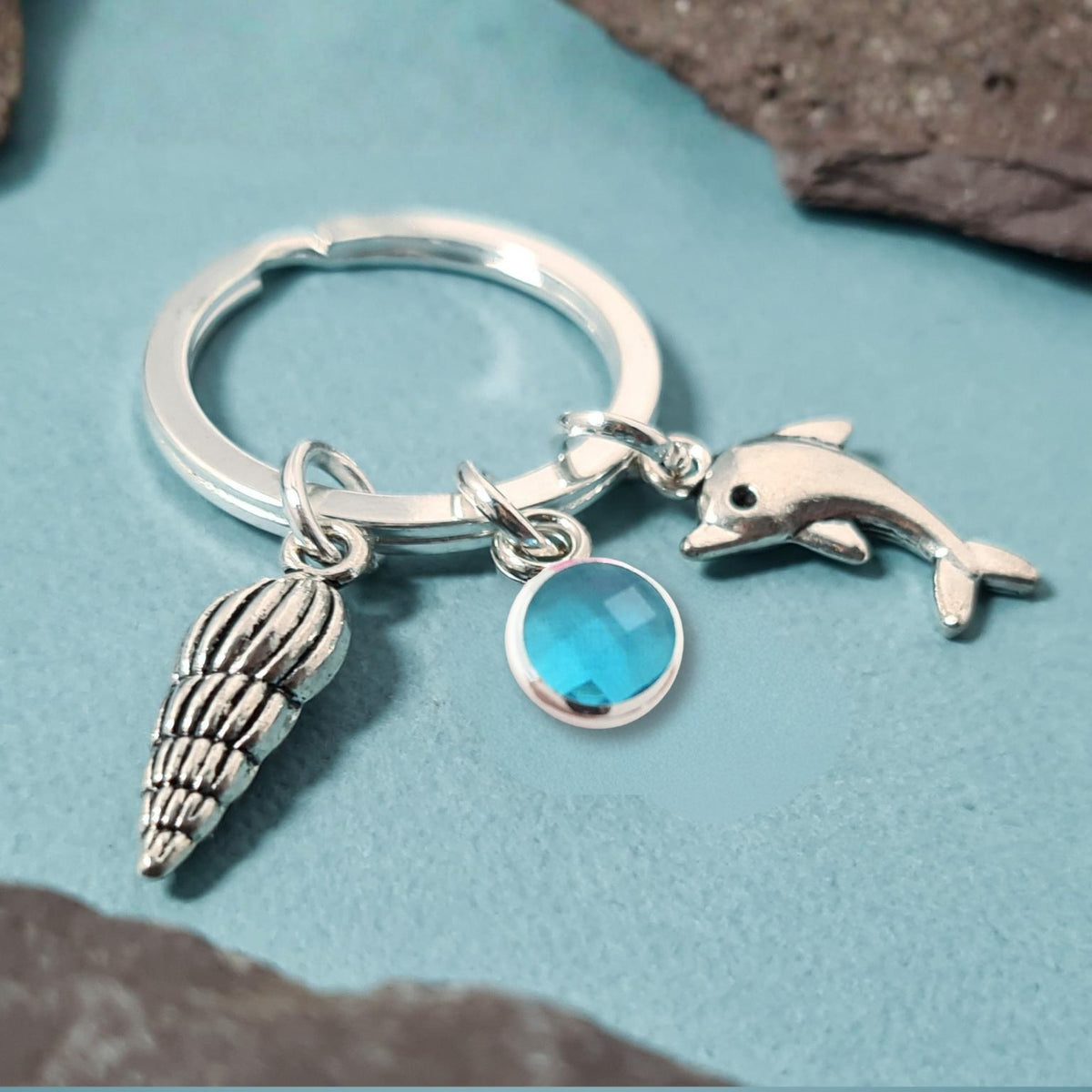 A silver keychain with a split ring, featuring charms of a seashell, a dolphin, and a faceted blue gemstone, displayed on a textured teal surface with dark rocks.