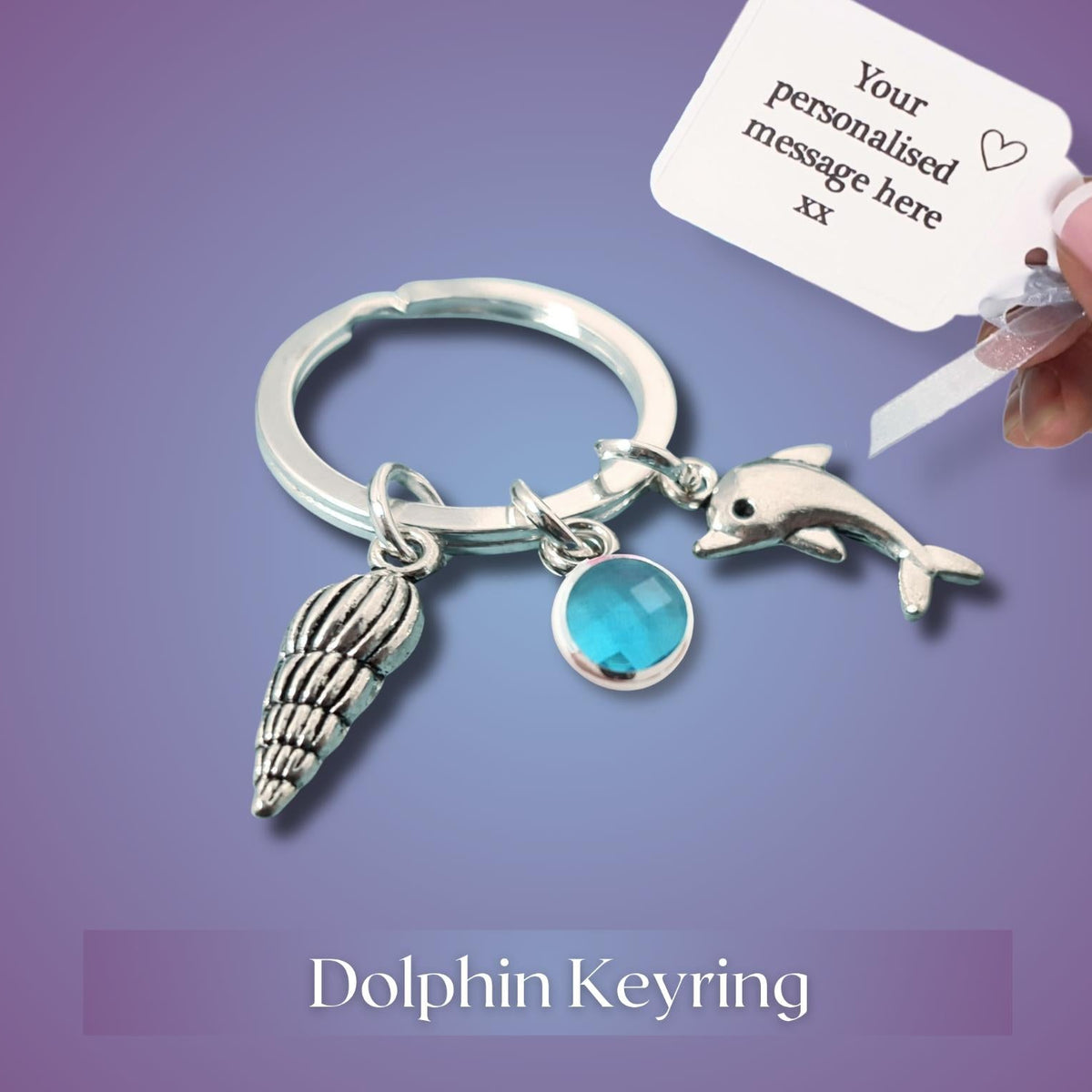 A silver dolphin keyring with a seashell charm, a blue oval stone charm, and a key tag being held by a hand, set against a purple gradient background with the text 'Dolphin Keyring' at the bottom.