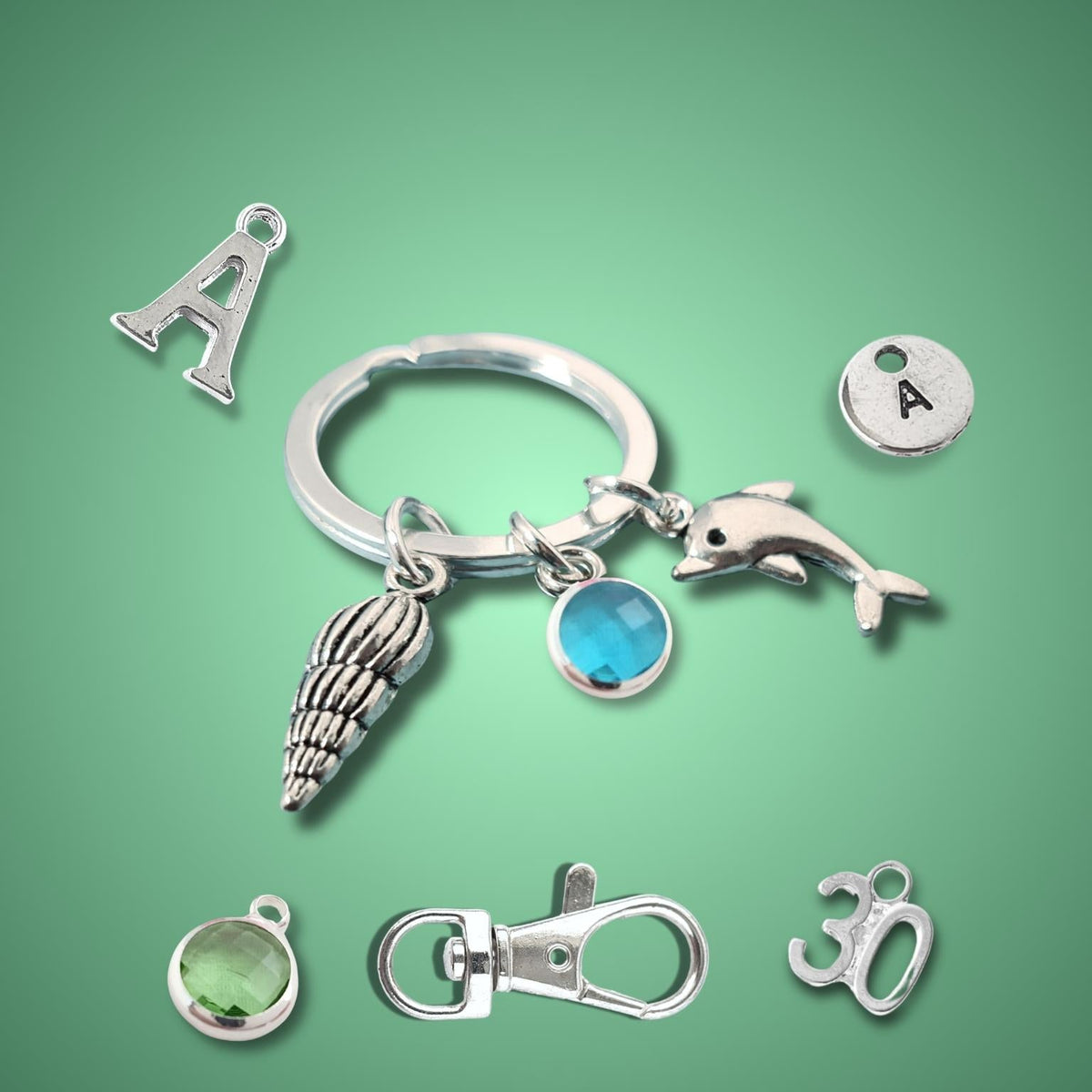 A set of silver-toned keychain charms arranged on a green gradient background, including an uppercase letter A, a dolphin charm, a shell charm, a round letter A disc, a blue oval gem, a green oval gem, a lobster clasp, and a 30th birthday charm.