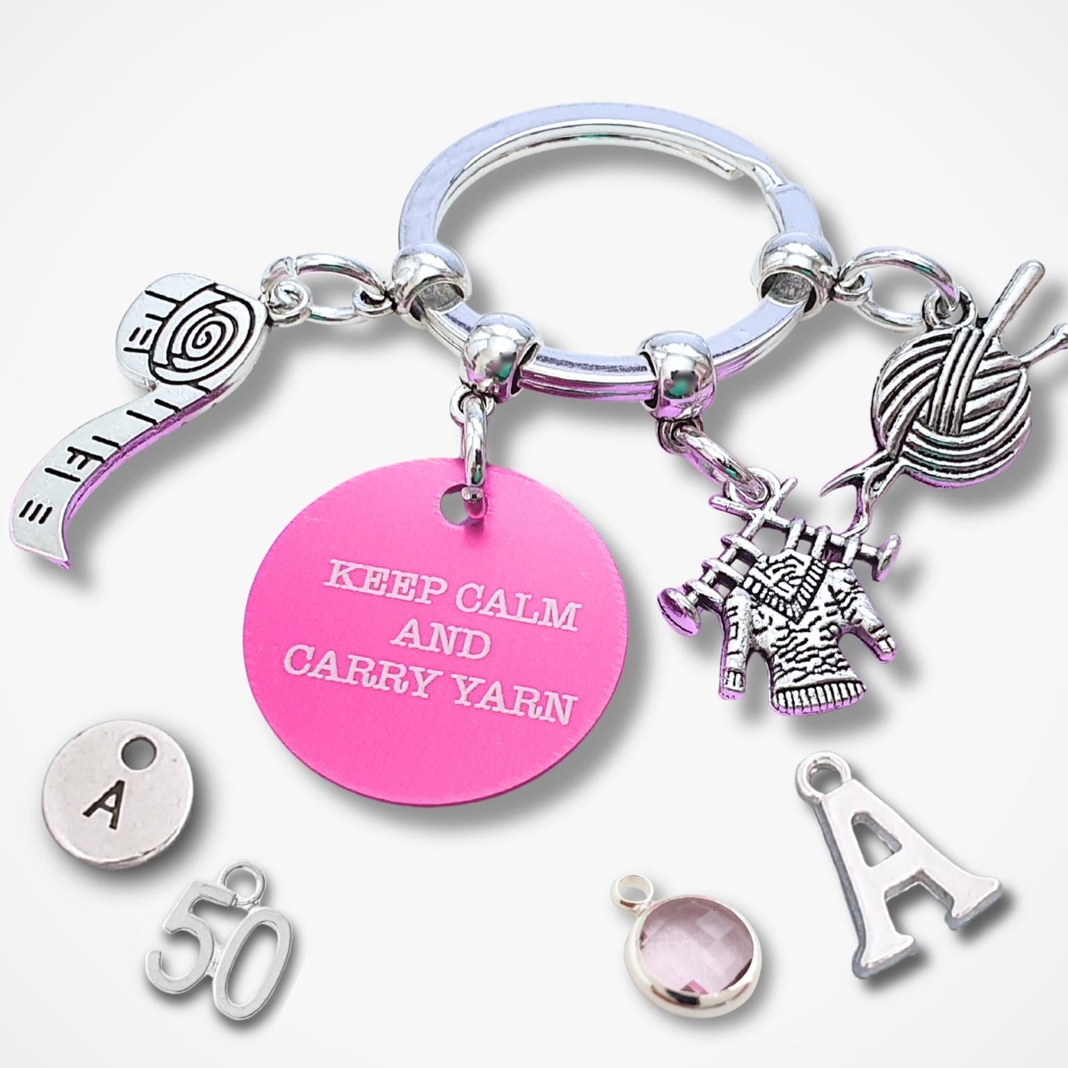 A silver-toned keychain with multiple charms, including a pink round charm engraved with 'KEEP CALM AND CARRY YARN', a knitting needle and yarn ball charm, and several loose letter and number charms on a light gray background.