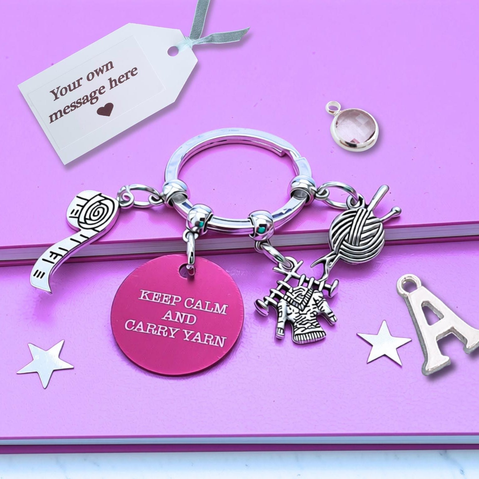 A silver keyring charm set displayed on a pink notebook. The set includes a round pink tag engraved with 'KEEP CALM AND CARRY YARN', a silver tape measure and yarn ball charm,  and a silver letter 'A' charm.