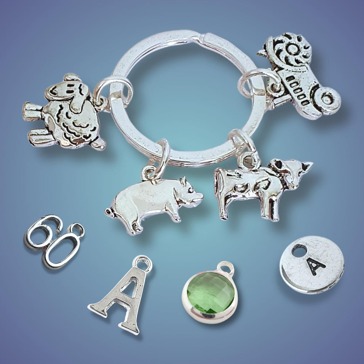 A farm theme keyring arranged on a blue gradient background. The keyring includes a sheep charm, a pig charm, a cow charm, a tractor charm, an 'A' charm, a round silver disc with the letter 'A', a green faceted gemstone bezel,