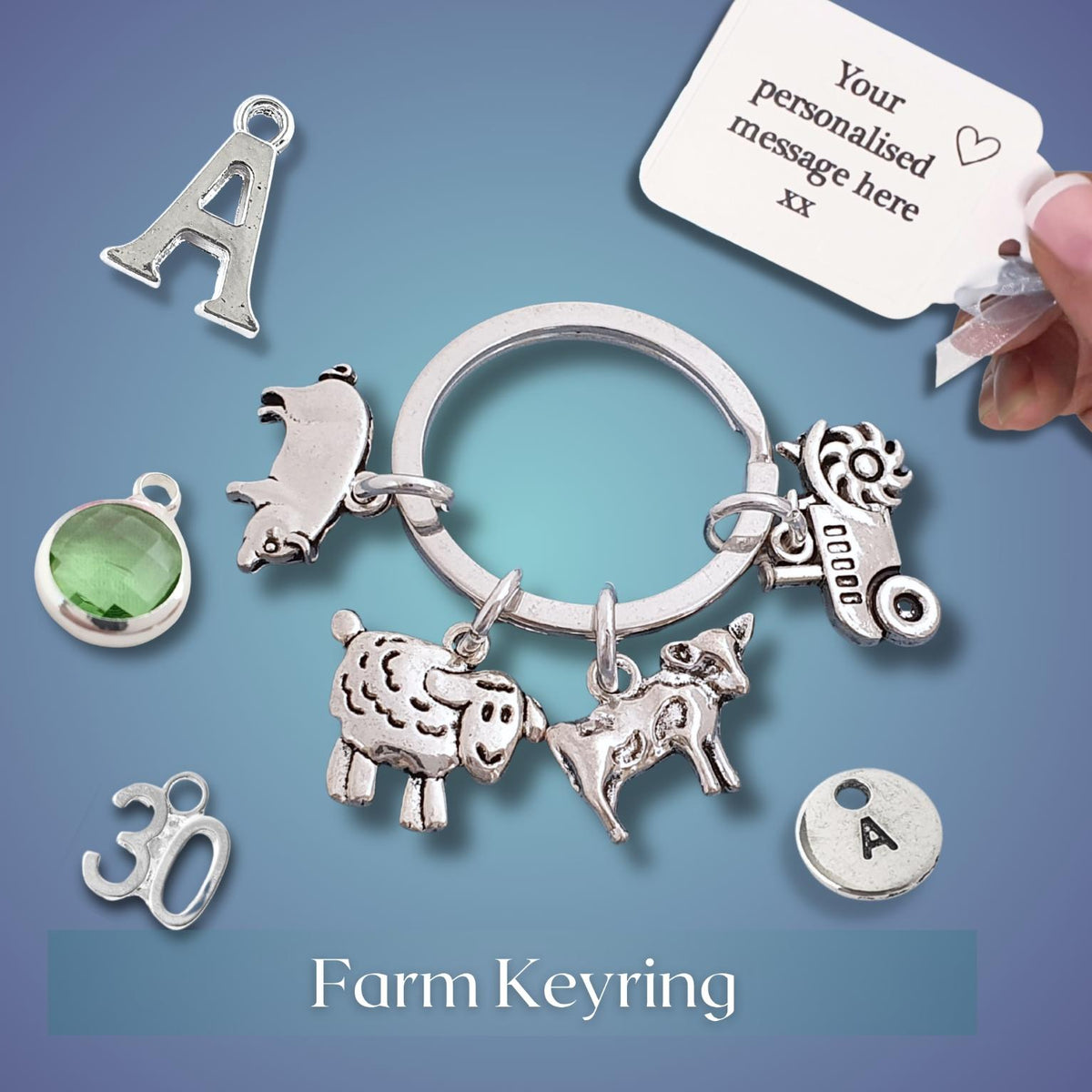 A silver-tone farm-themed keyring charm set displayed on a blue gradient background, with a hand holding a white personalization tag. The keyring includes charms shaped like a pig, sheep, cow, tractor, along with separate letter and symbol charms.