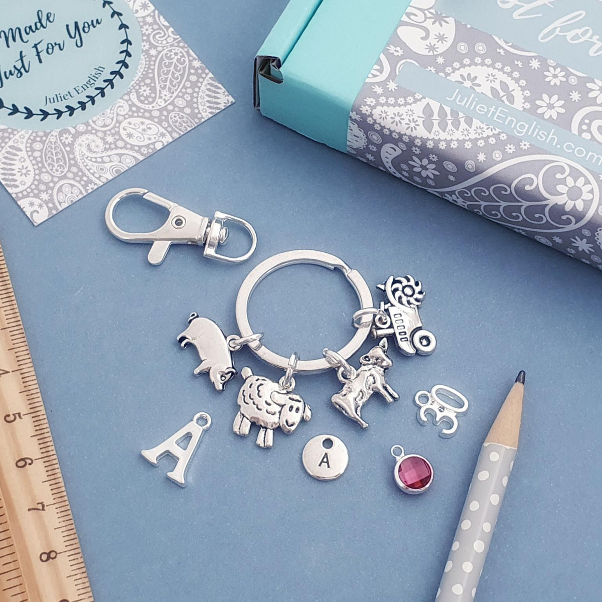 Silver charm keyring with multiple animal and letter charms, displayed on a blue surface with a turquoise gift box, wooden ruler, pencil, and a small card.
