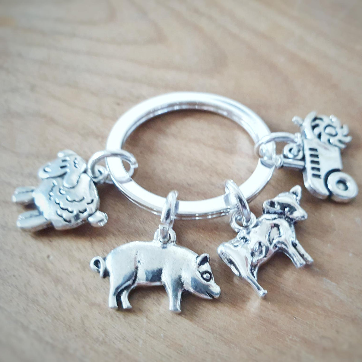 A silver-colored metal key ring charm set featuring four small farm-themed animal and vehicle charms, including a sheep, pig, cow, and tractor, displayed on a light wood surface.
