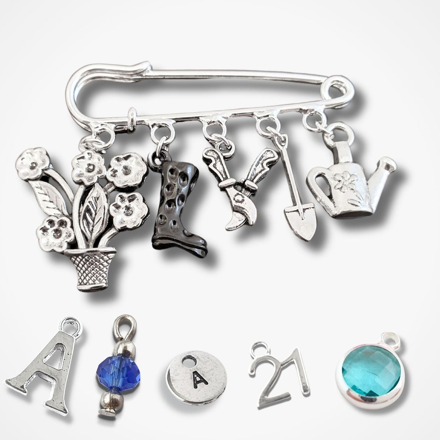 A silver safety pin brooch adorned with various gardening and outdoor-themed charms, including a flower pot, boots, shears, shovel, watering can, and a letter 'A' charm, displayed on a light gray background.