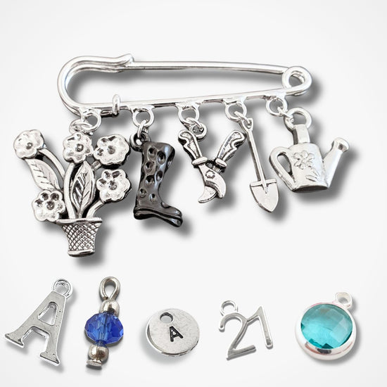 A silver safety pin brooch adorned with various gardening and outdoor-themed charms, including a flower pot, boots, shears, shovel, watering can, and a letter 'A' charm, displayed on a light gray background.