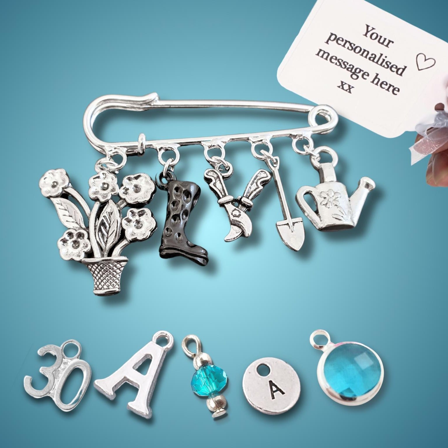 A silver-tone personalized garden-themed brooch pin with multiple charms, shown with a hand holding a white sample tag that reads 'Your personalised message here xx'. The charms include a flower pot, flowers, a black boot, gardening tools, a watering can, and letter 'A' charms, with other silver charms displayed below on a blue gradient background.