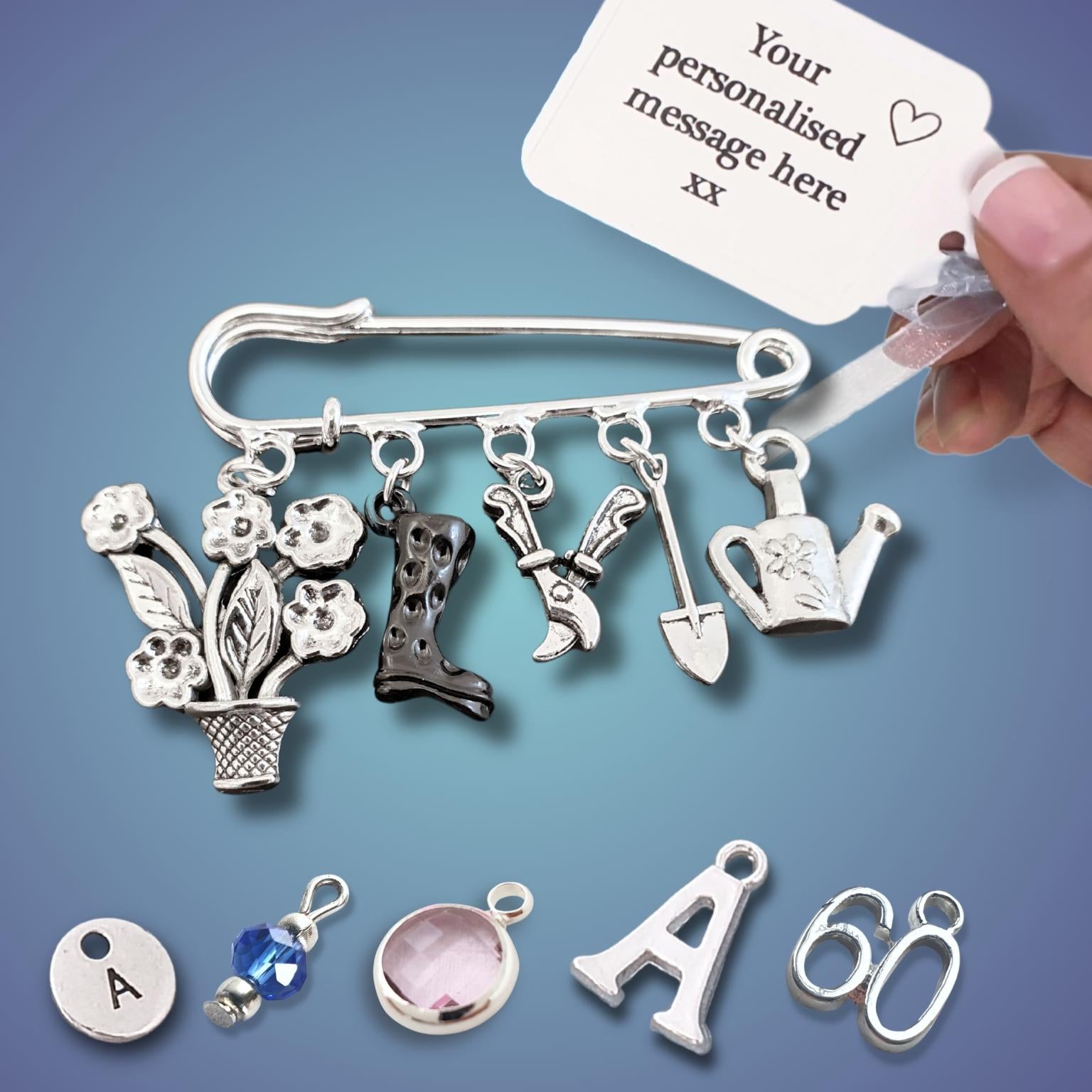A silver-toned safety pin charm brooch with multiple gardening-themed charms including a flower pot with flowers, a cowboy boot, gardening tools, and a watering can, shown with a white personalization tag and a hand holding it. Additional small charm options are displayed below on a blue gradient background.