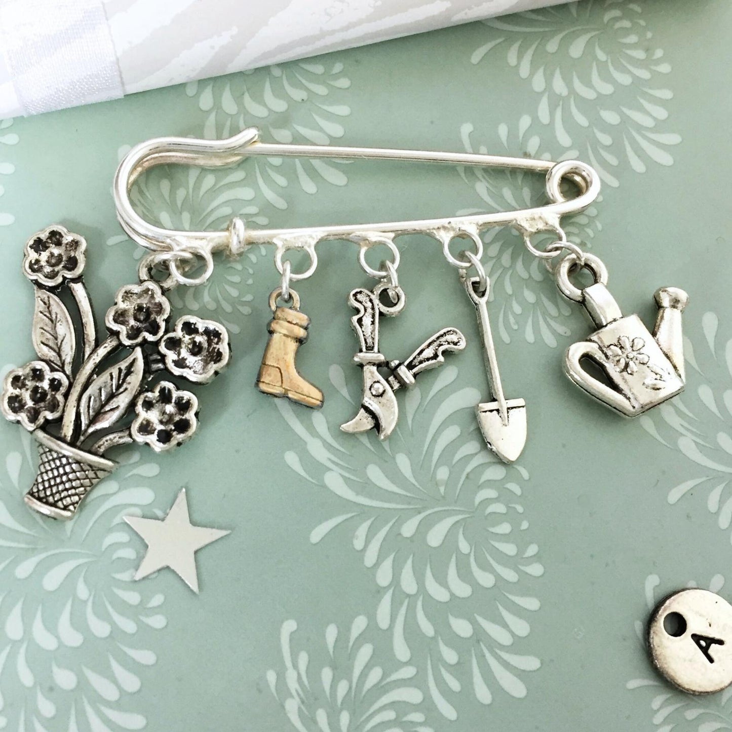 A silver safety pin brooch with five gardening-themed charms, including a flower pot, boots, gardening tools, and a watering can, displayed on a light green patterned background.