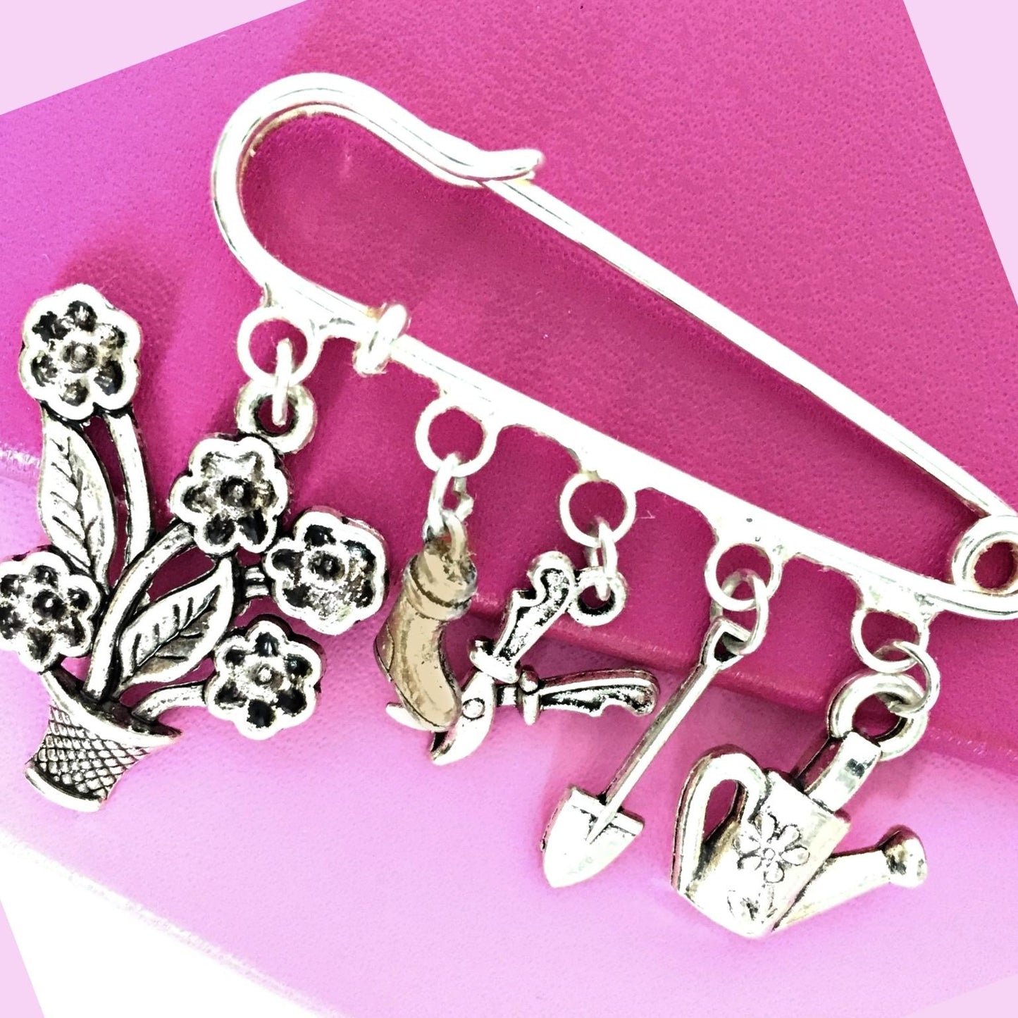 A silver safety pin brooch decorated with dangling charms of a potted flower, a boot, a garden tool, and a watering can, displayed on a pink and purple background.