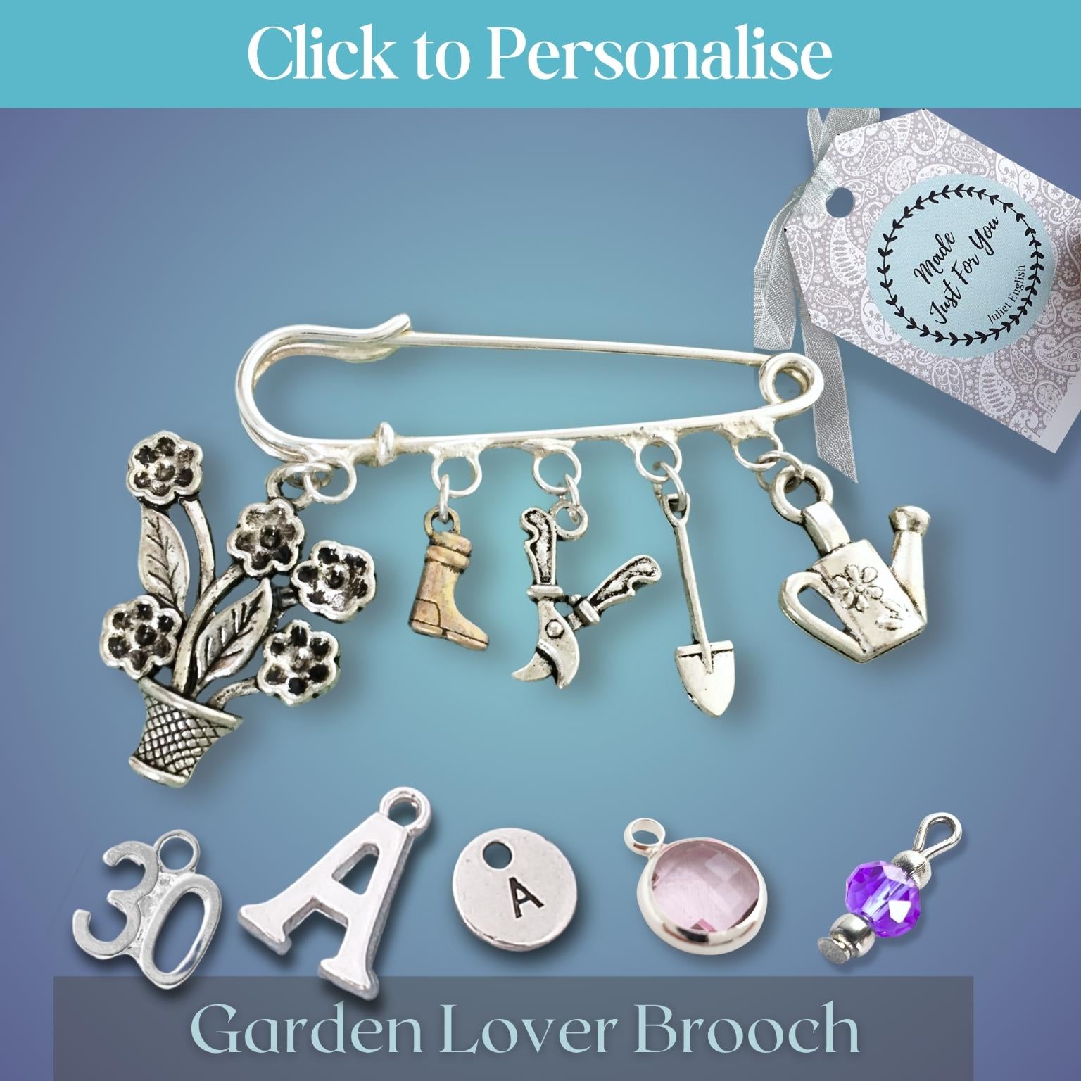 A silver-tone garden-themed brooch shaped like a safety pin with dangling charms including flowers, gardening tools, a watering can, boots, and a pot, shown on a blue gradient background with a hand holding a personalized tag.