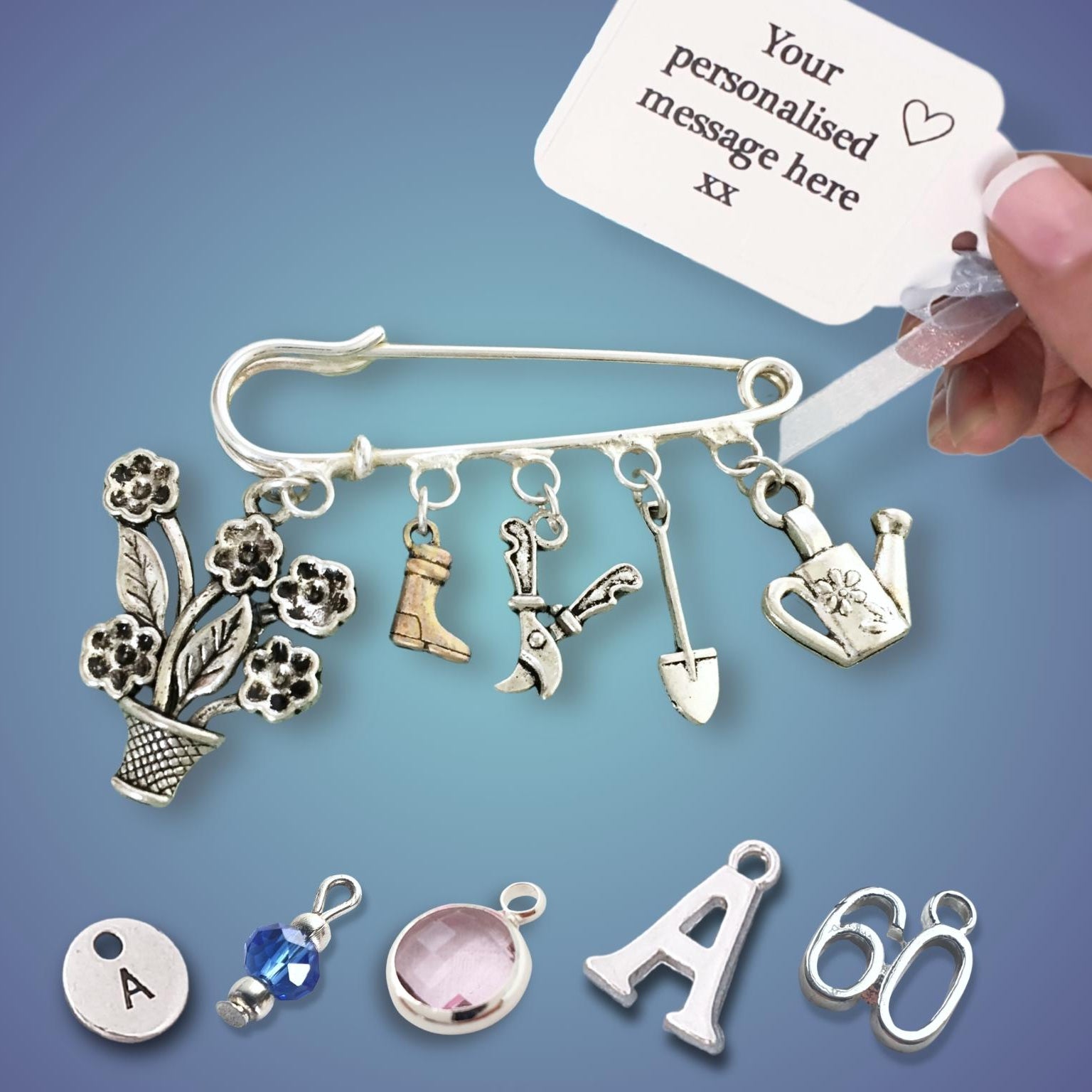 A hand holds a white tag that reads 'Your personalised message here xx' above a silver safety pin charm set with floral, boot, gardening, watering can, and letter charms. Smaller silver charms are arranged below on a blue gradient background.