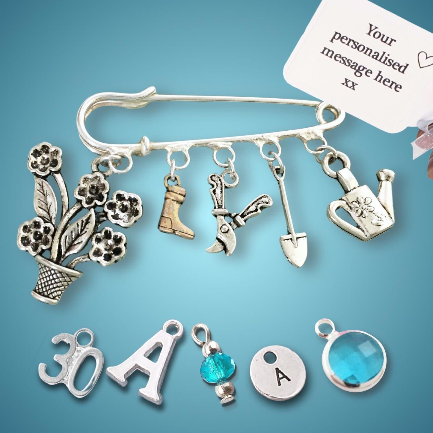 A silver-tone safety pin charm bar with gardening-themed charms including a flower pot with flowers, boots, gardening tools, and a watering can, shown on a blue gradient background. A hand holds a white sample tag next to it, and additional customizable charms are displayed below.