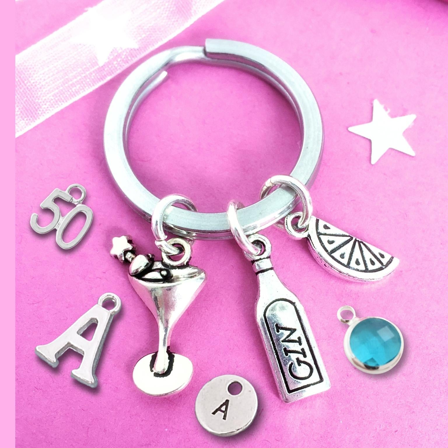 A silver-tone keychain with multiple charms, including a martini glass, a bottle labeled 'GIN', a lemon slice, a blue gemstone, and letter 'A' charms, displayed on a pink background.