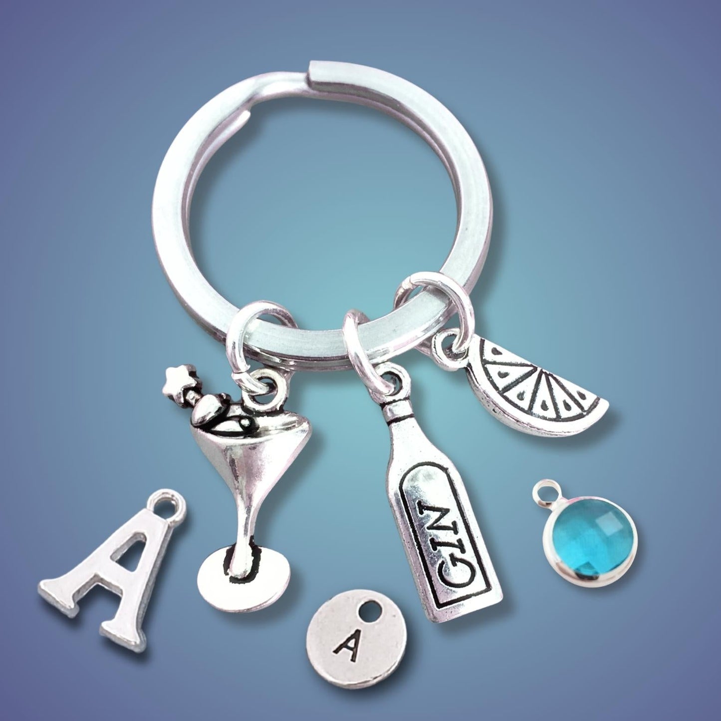 A silver-tone keychain with multiple cocktail-themed charms, including a martini glass, a bottle labeled 'GIN', a slice of lime, a letter 'A' charm, and a round letter 'A' disc, shown on a blue gradient background.